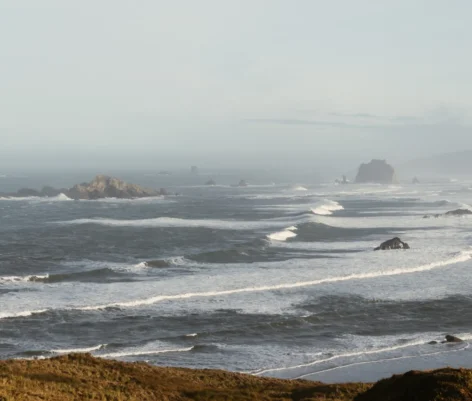 Vista panorámica de un océano con olas y rocas, bajo un cielo nublado y una atmósfera serena.
