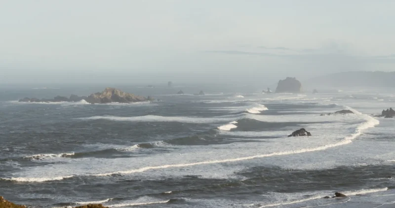 Vista panorámica de un océano con olas y rocas, bajo un cielo nublado y una atmósfera serena.