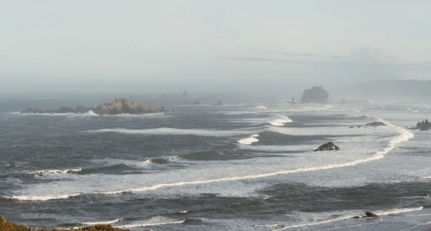 Vista panorámica de un océano con olas y rocas, bajo un cielo nublado y una atmósfera serena.