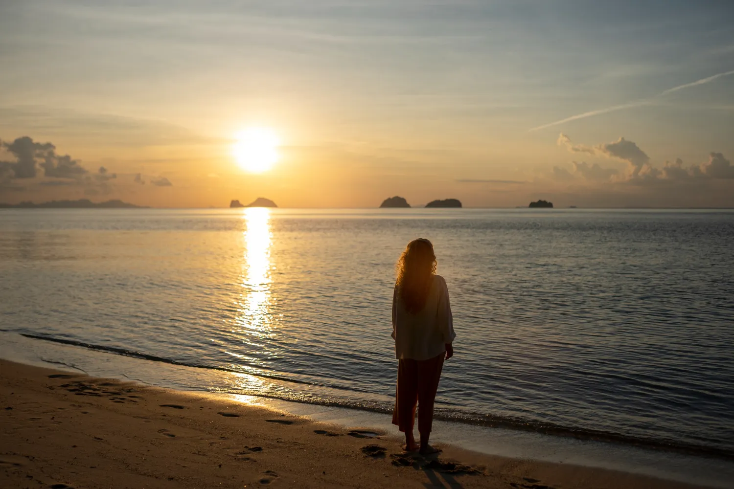 Mujer de pie en la playa contemplando un atardecer sobre el mar, con islas a lo lejos y reflejos en el agua.